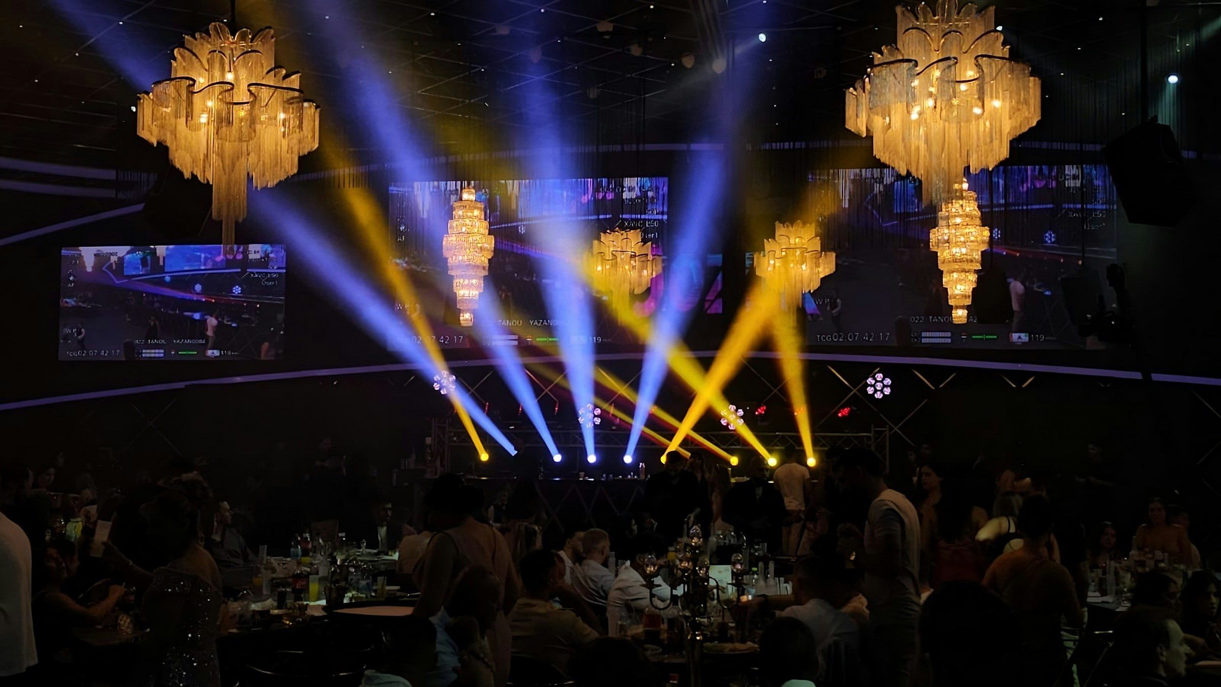 Beams of coloured light cutting through a darkened event hall beneath chandeliers